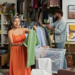 Young smiling woman looking at green blouse on hanger while standing in thrift shop and choosing new casual apparel