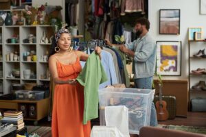 Young smiling woman looking at green blouse on hanger while standing in thrift shop and choosing new casual apparel