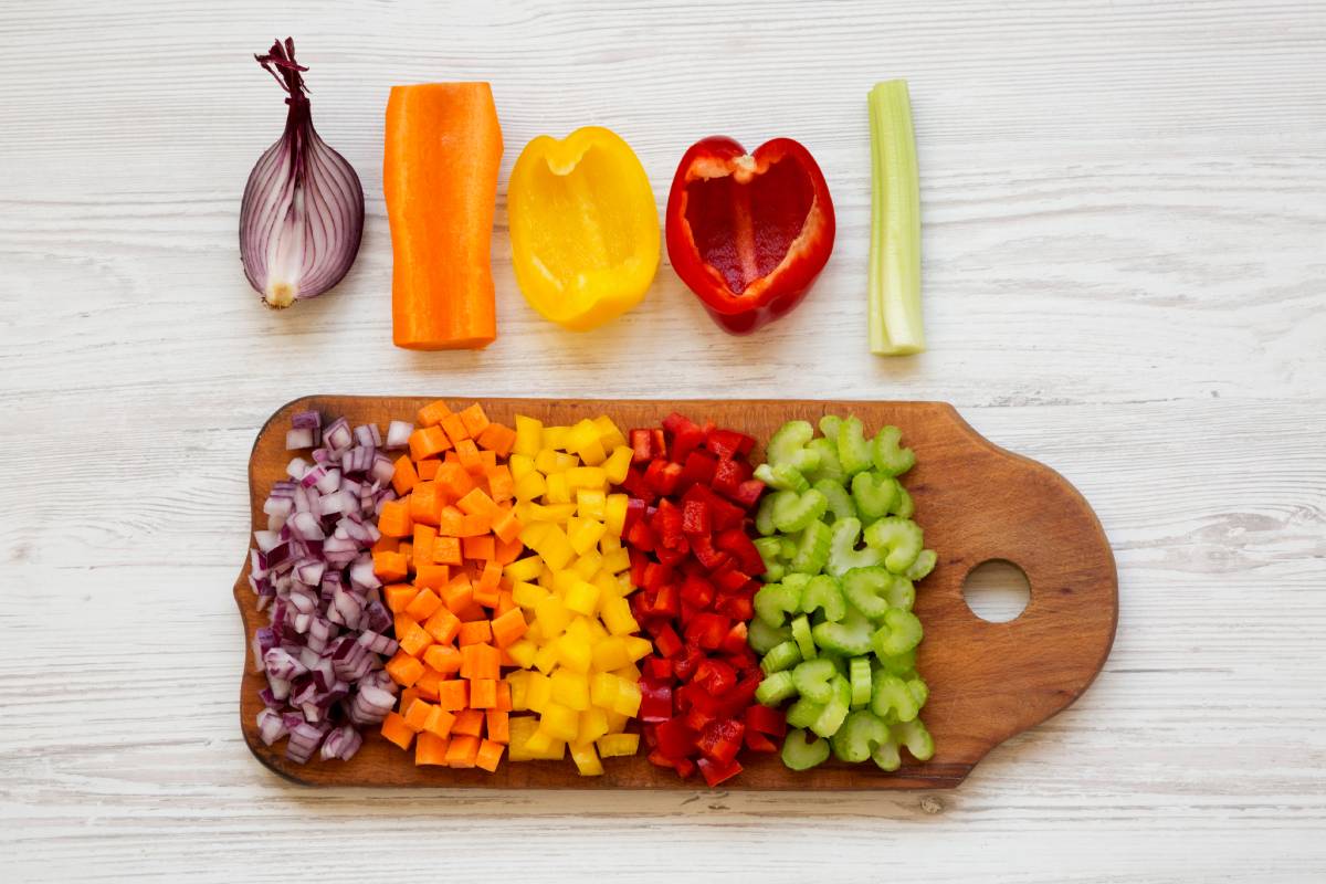 Chopped fresh vegetables (carrot, celery, onion, colored peppers) arranged on cutting board on a white wooden surface, top view. Flat lay, overhead, from above.