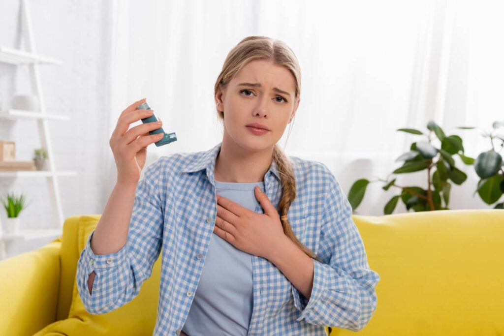 Displeased woman with allergy reaction holding inhaler on couch