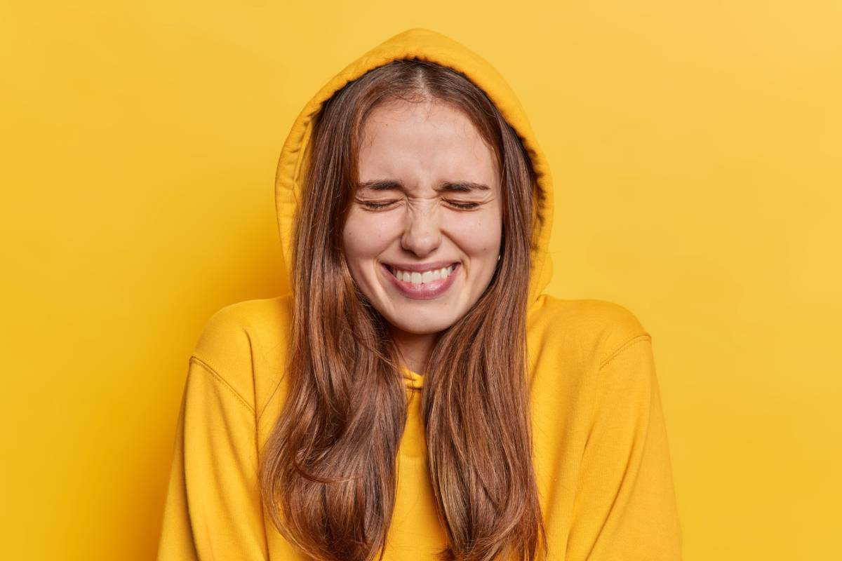 Overjoyed dark haired young woman keeps eyes closed giggles positively enjoys candid moment of fun wears hoodie with hood on head expresses positive emotions isolated over yellow background.