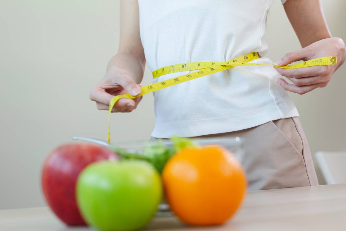 woman measures her waist size with tape measure with fruit on table foreground. female checking waist size eat fruits and vegetables, healthy care body conacept.