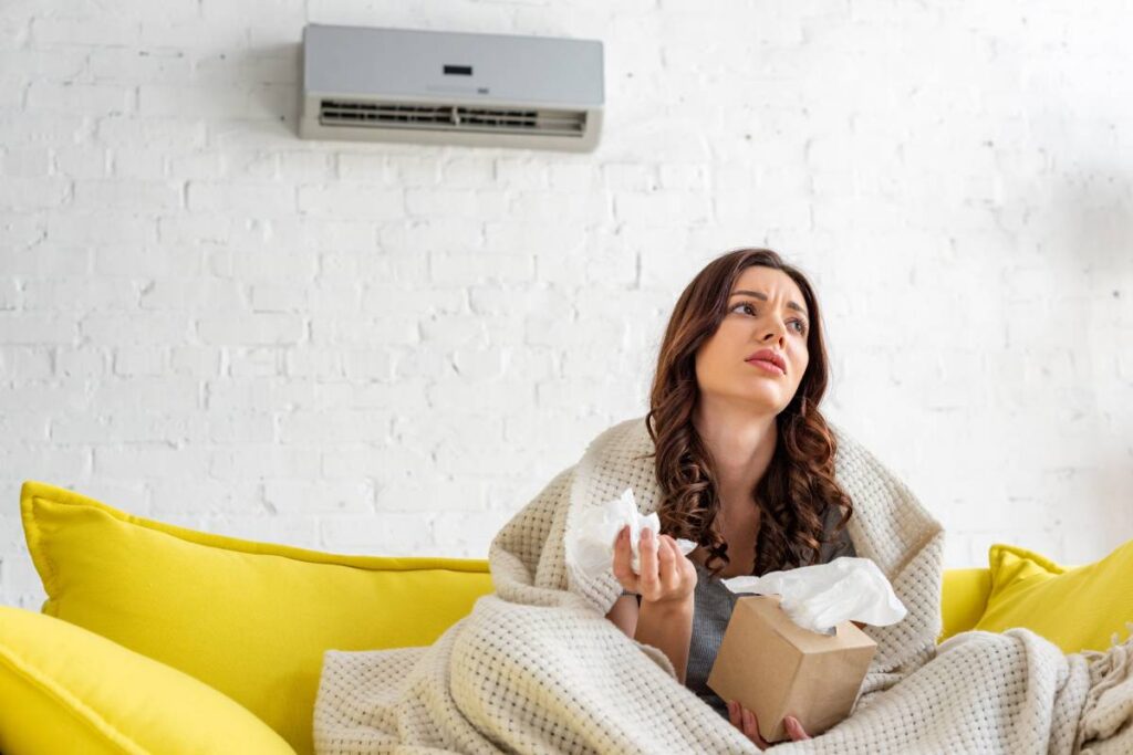 pretty sick woman holding paper napkins while warming under blanket under air conditioner