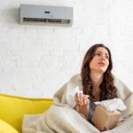 pretty sick woman holding paper napkins while warming under blanket under air conditioner