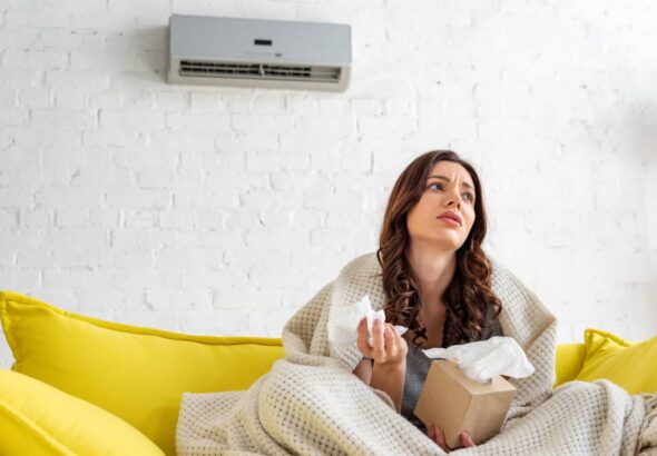 pretty sick woman holding paper napkins while warming under blanket under air conditioner