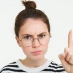 Portrait of young woman, tutor in glasses, pointing finger up and frowning, showing smth important, standing over white background