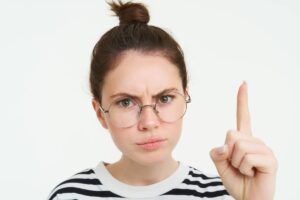 Portrait of young woman, tutor in glasses, pointing finger up and frowning, showing smth important, standing over white background