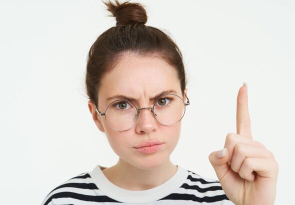Portrait of young woman, tutor in glasses, pointing finger up and frowning, showing smth important, standing over white background